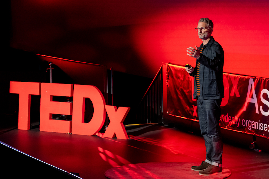 Man speaking at TEDx talk on stage red lights