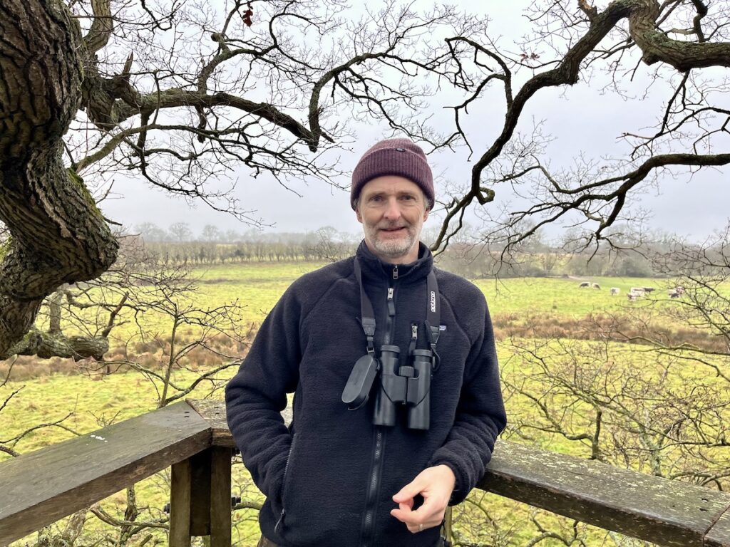 man with beard and hat in treehouse binoculars around neck