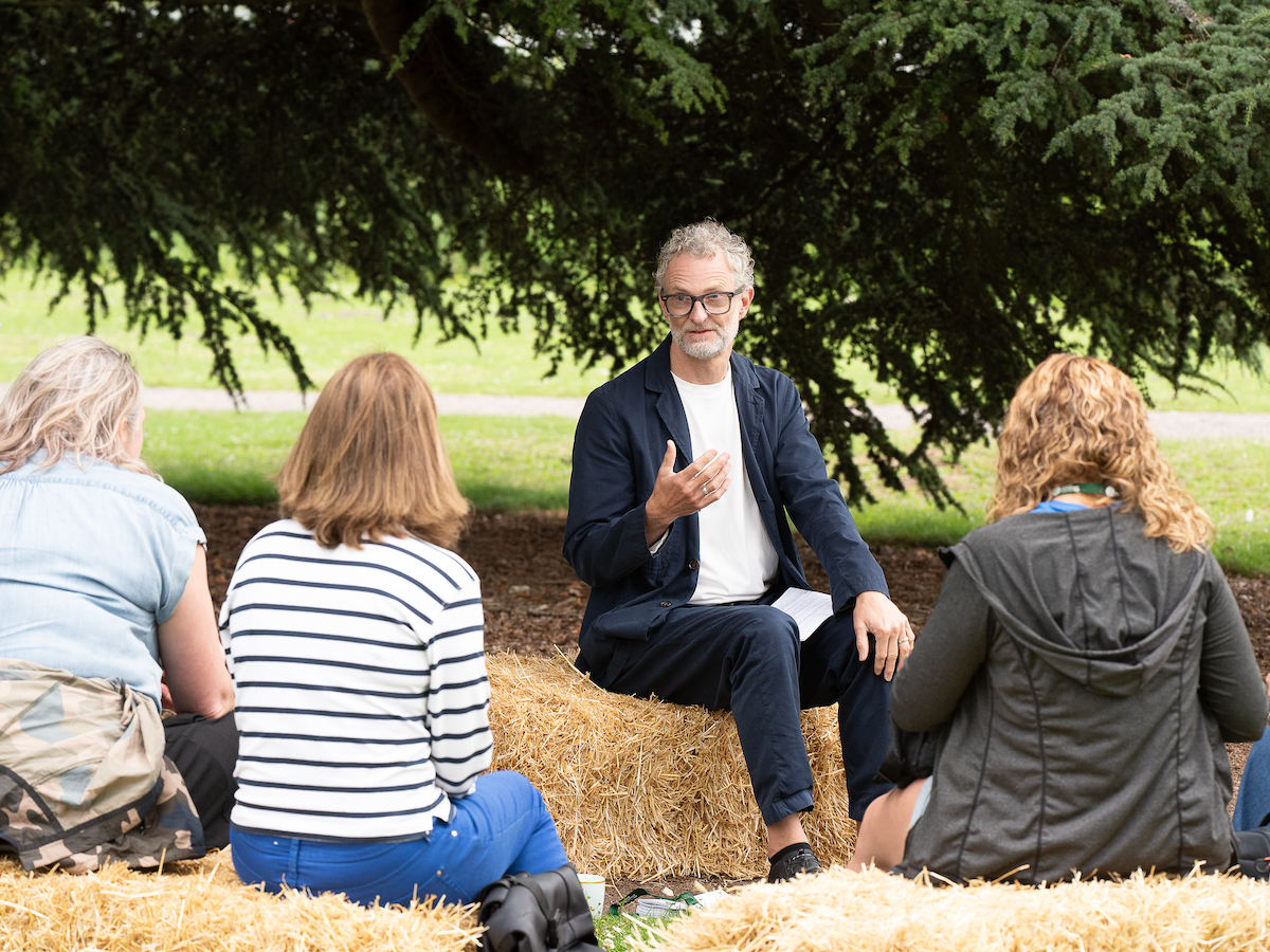 Man in glasses speaking to group sitting on straw bales outdoors